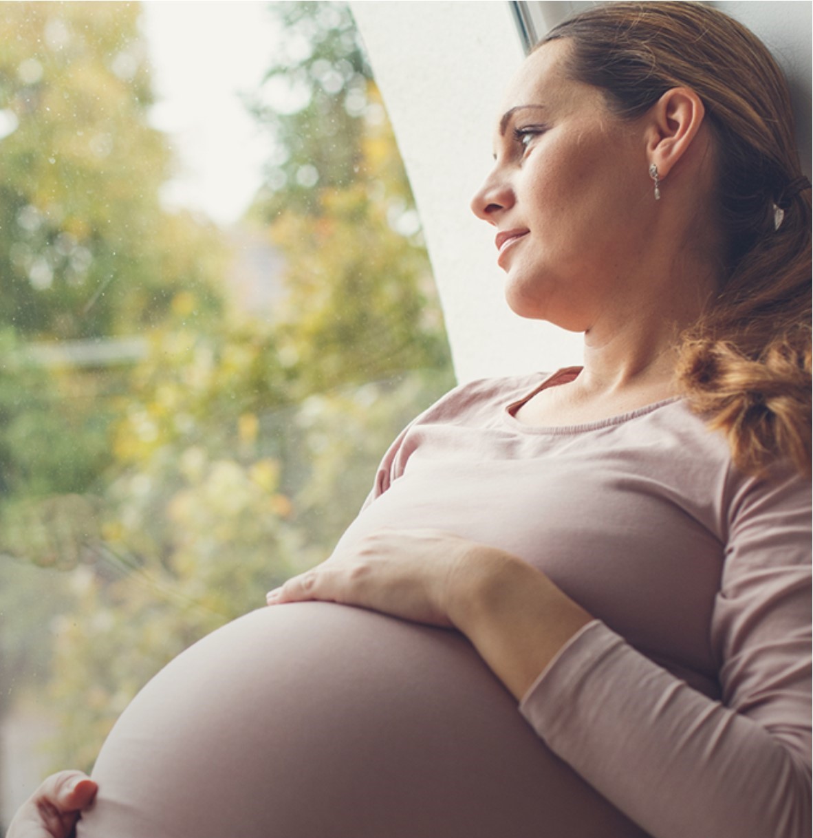 Pregnant woman looking out the window holding her belly